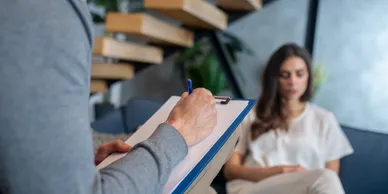 A person writing on paper with another person sitting in the background.