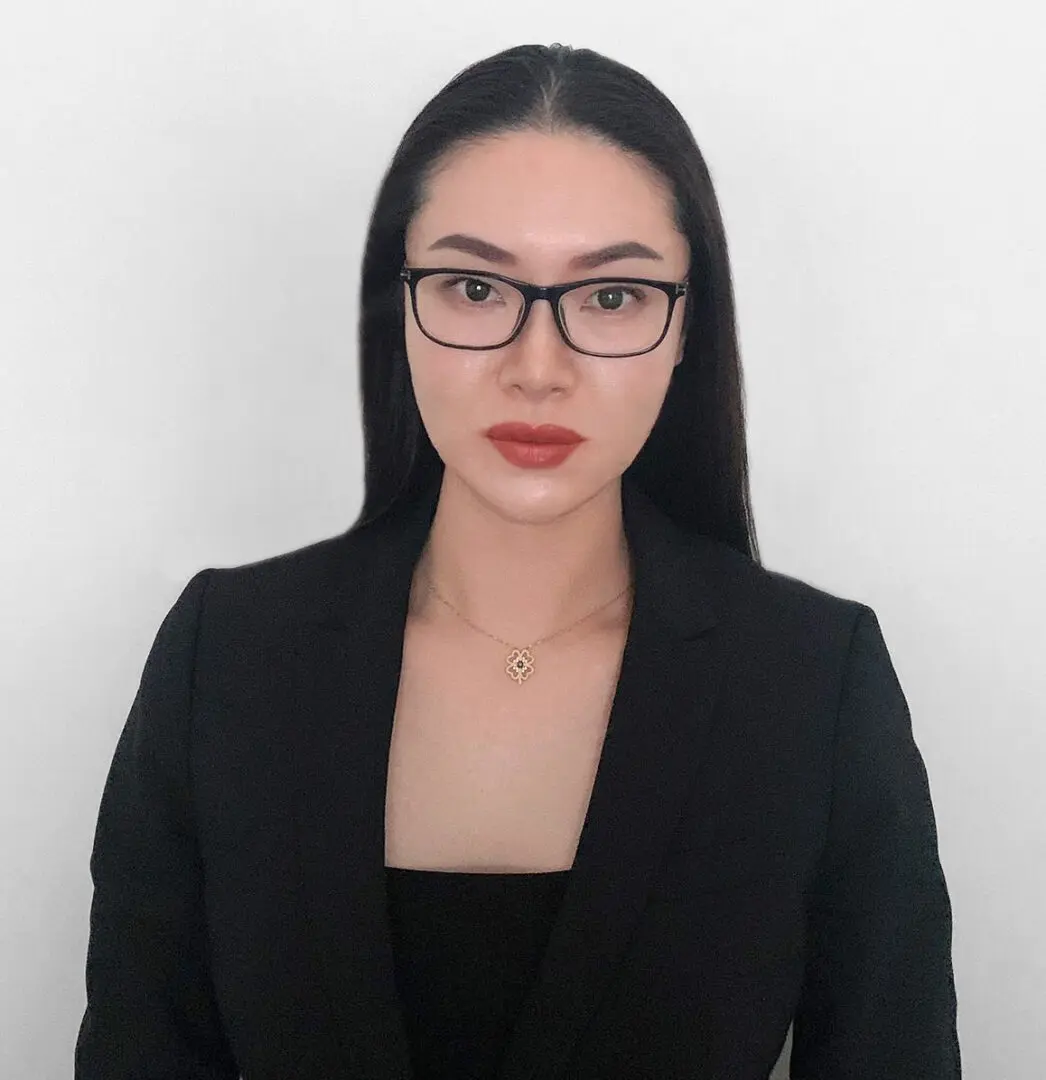 A woman in black jacket and glasses standing next to wall.